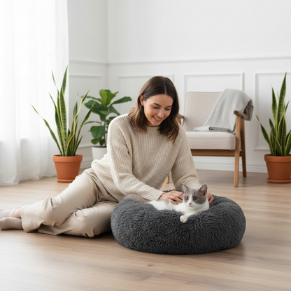 Woman sitting on the floor with a cat on a fluffy gray pet bed in a bright room with plants.